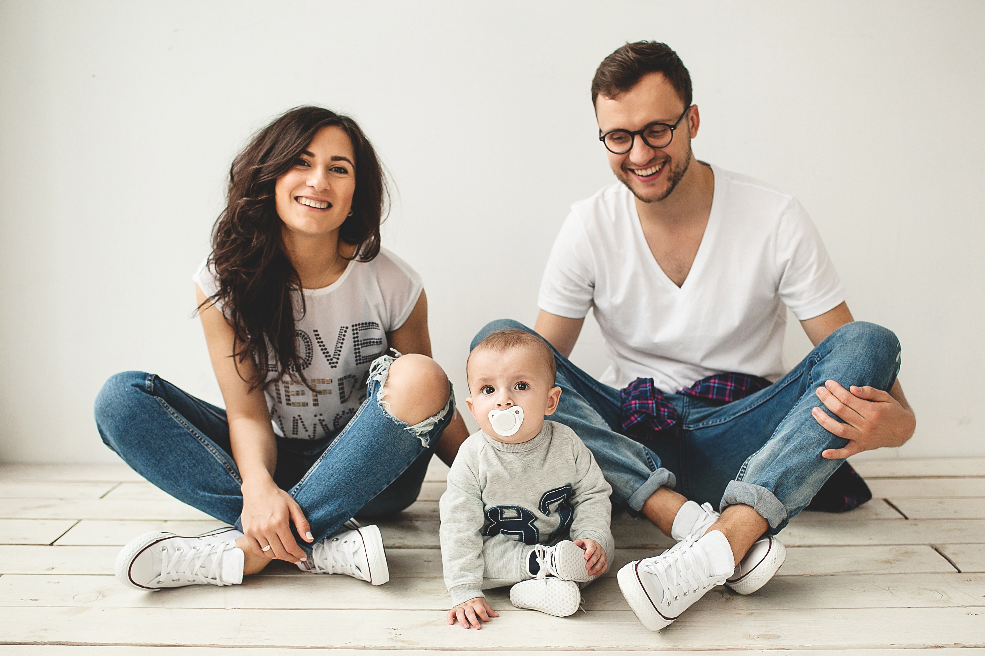 Young hipster father, mother and cute baby boy sitting on rustic wooden floor over white background